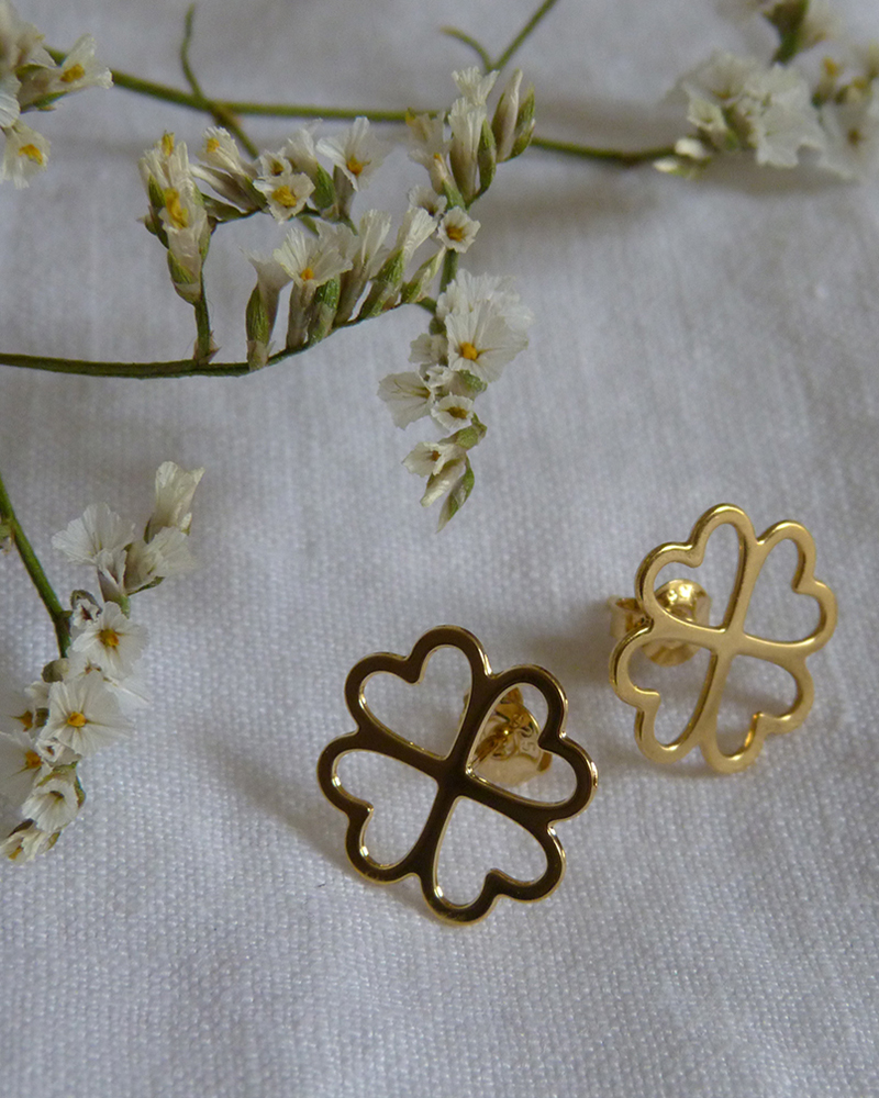 Puces d'oreilles trèfle à quatre cœurs dorées posées sur un tissu blanc, avec de petites fleurs séchées