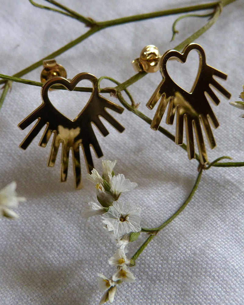 Puces d'oreilles cœur rayonnant doré accrochées à une branche de petites fleurs blanches, sur tissu clair